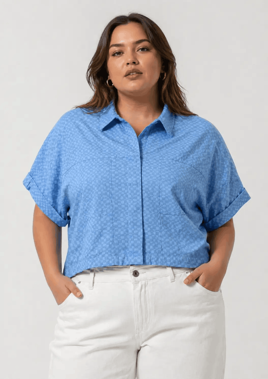Woman wearing a blue patterned shirt and white pants on a plain background