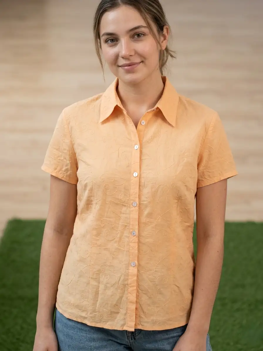 Woman wearing an orange shirt standing outdoors with a wooden wall and green grass in the background