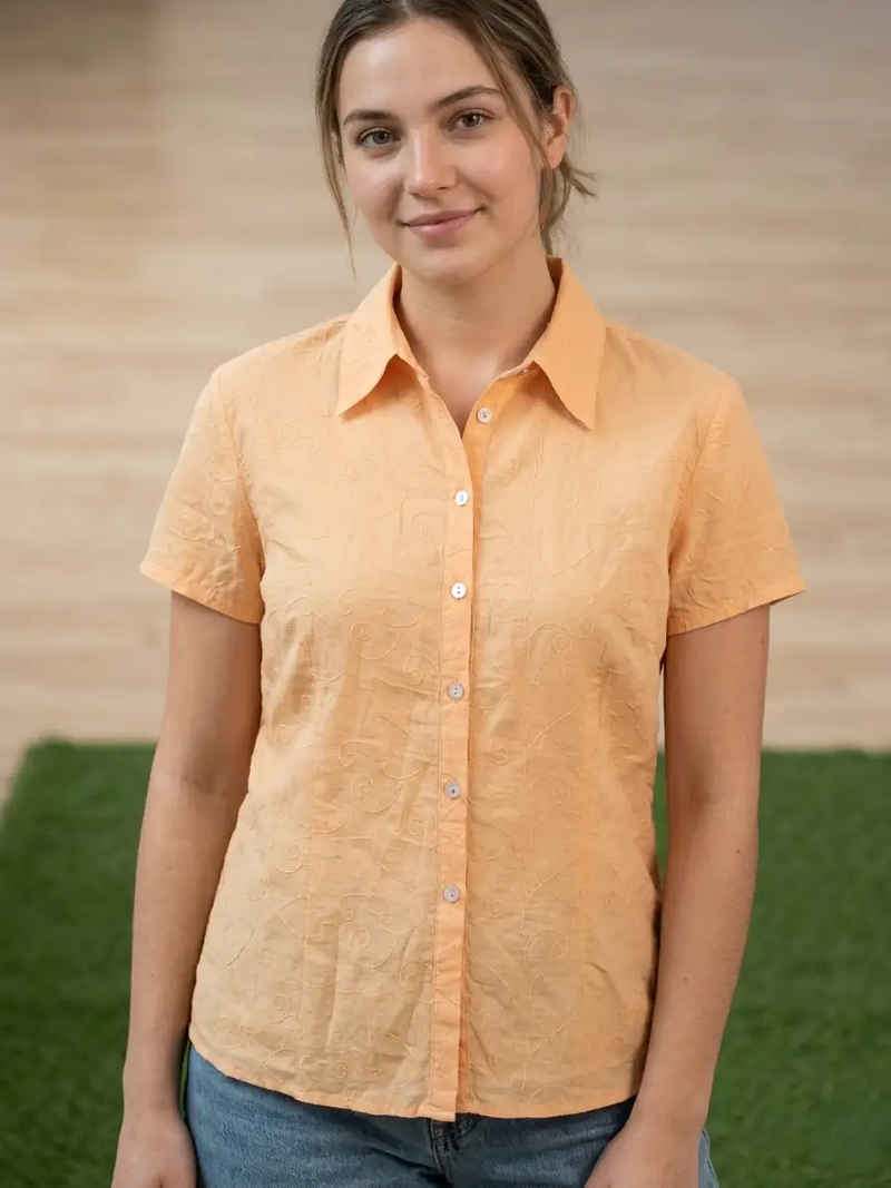 Woman wearing an orange shirt standing outdoors with a wooden wall and green grass in the background
