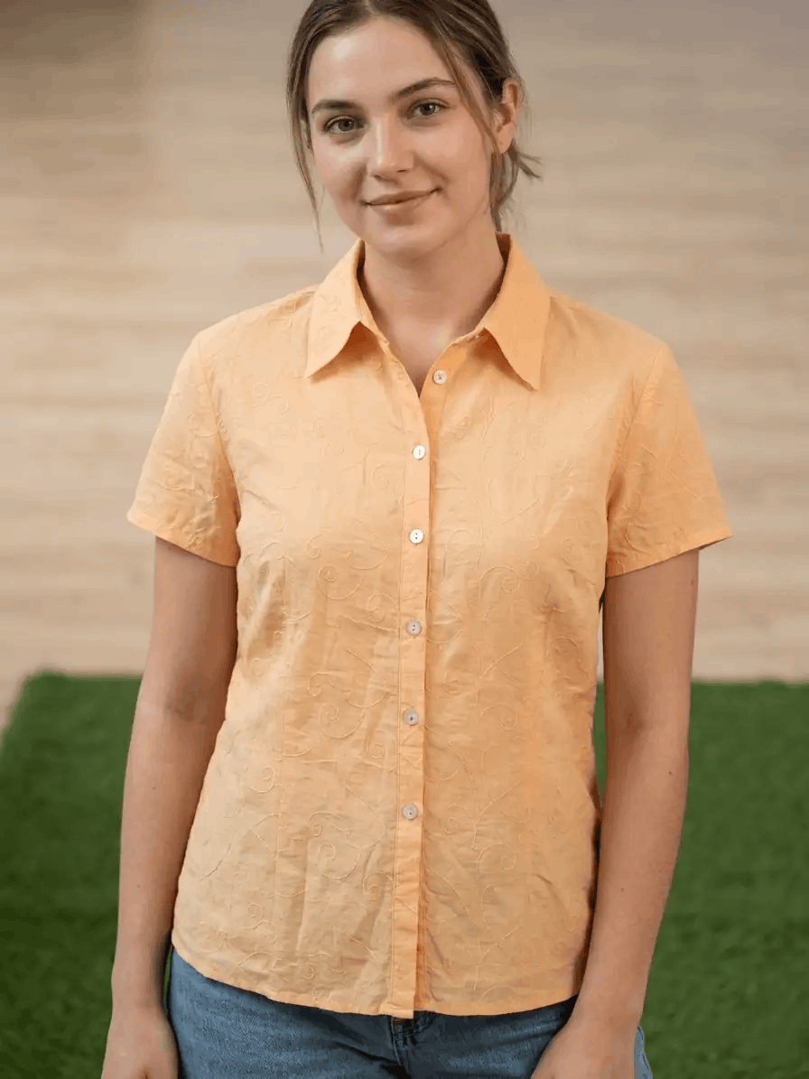 Woman wearing an orange shirt standing outdoors with a wooden wall and green grass in the background