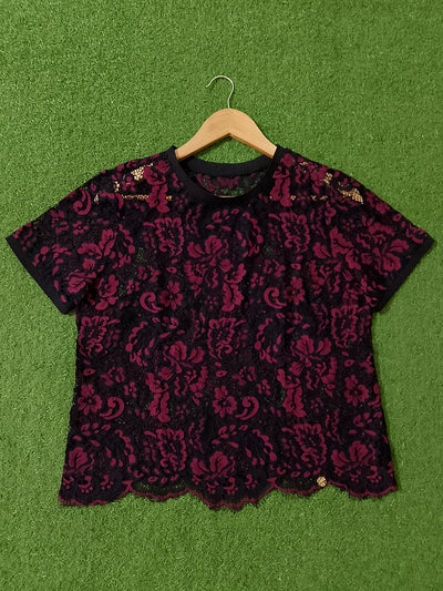 A black and deep magenta lace short-sleeve top with a ruffled neckline, displayed flat on a green artificial turf background with a wooden hanger.