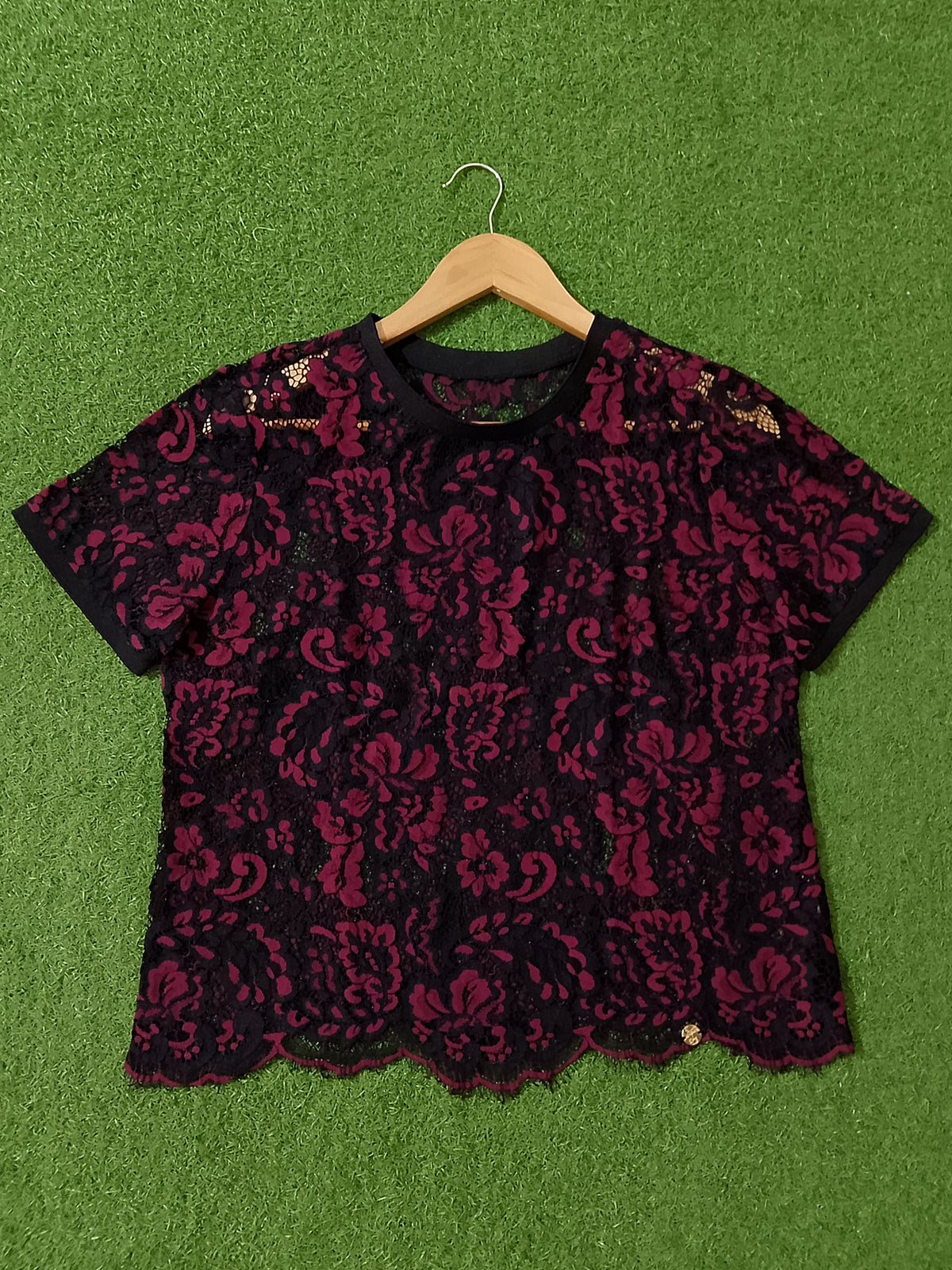A black and deep magenta lace short-sleeve top with a ruffled neckline, displayed flat on a green artificial turf background with a wooden hanger.